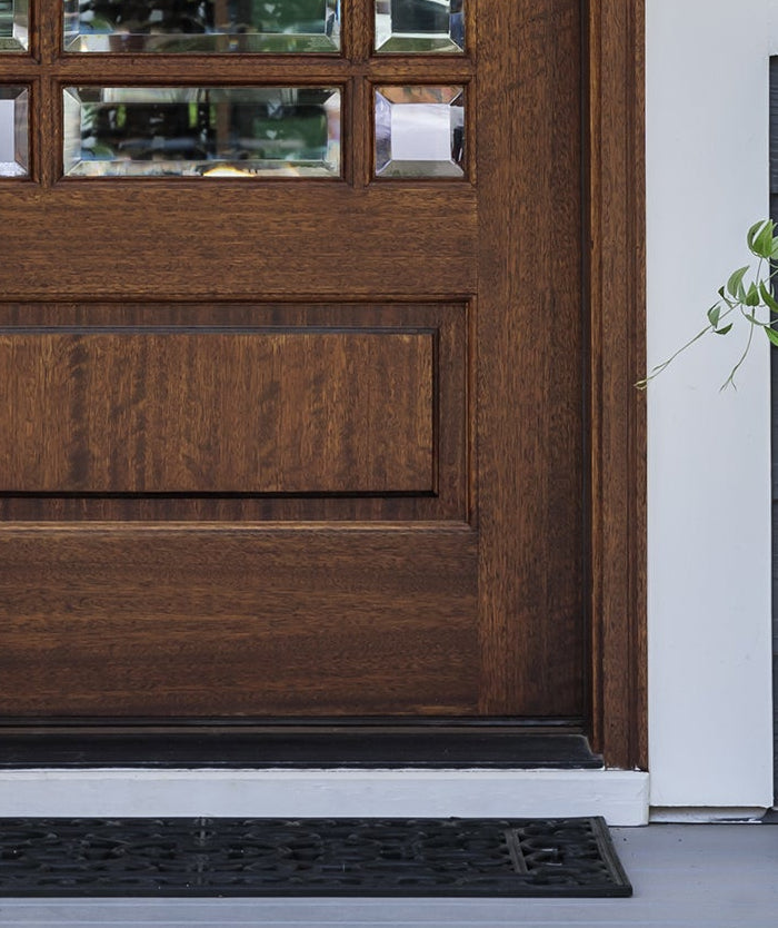 A front porch with a CYC tapered corten planter, a black doormat, a white lantern, and a large brown planter with green trailing plants on a gray porch floor.