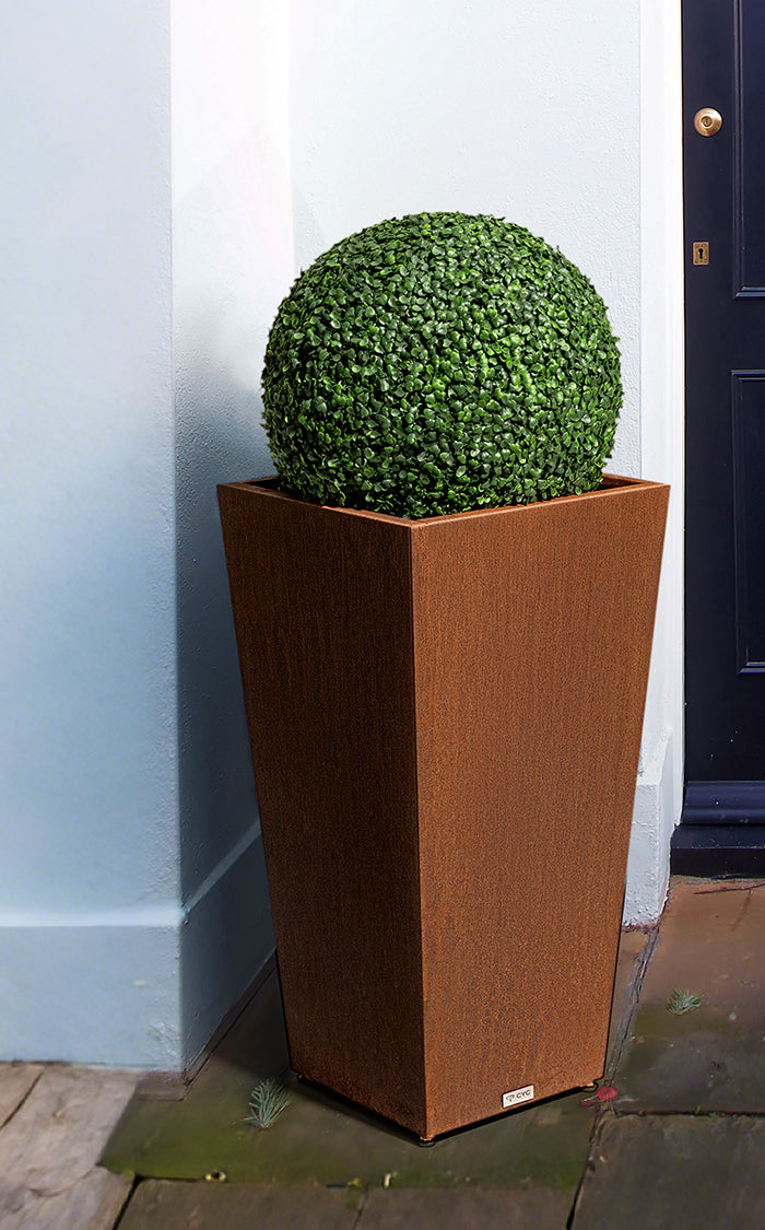 Tall tapered rust-colored planter with a spherical green topiary beside a navy front door and pale blue wall.