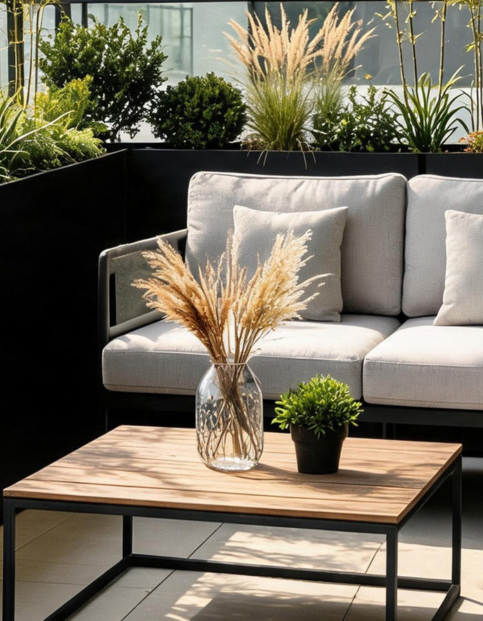Black rectangular planters with ornamental grasses and shrubs surrounding a cushioned sofa on a city balcony.