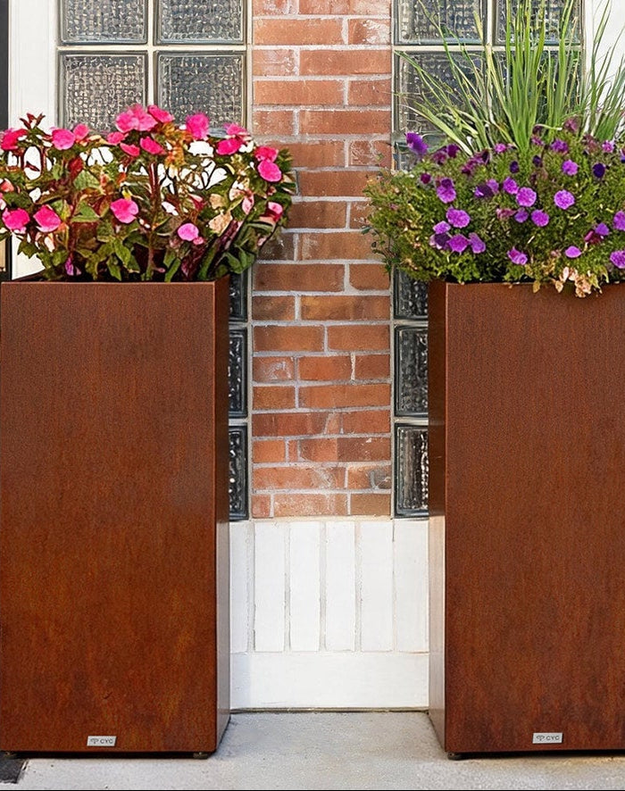 Two CYC tall rectangular rusted-corrigan planters filled with pink, purple, and green flowers, placed on either side of a front door with black doors and a brick column.